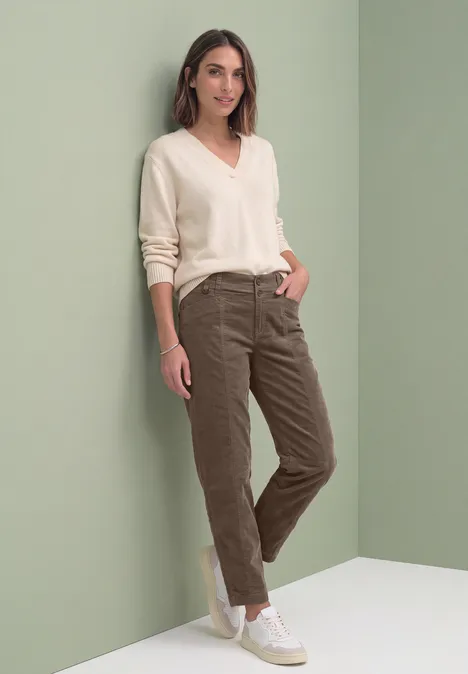 Barrel Leg Hose aus Cord im Casual Fit velvet mocha Frau lehnt lässig an hellgrüner Wand, trägt hellen V-Pullover, olivgrüne Hose und weiße Sneaker und lächelt.