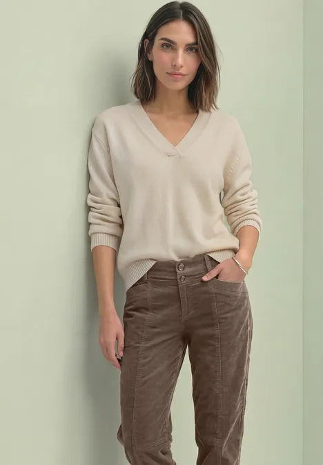 Barrel Leg Hose aus Cord im Casual Fit velvet mocha Frau in beigem V-Pullover und braunen Cordhosen steht vor neutralem Hintergrund, Hand in Tasche.