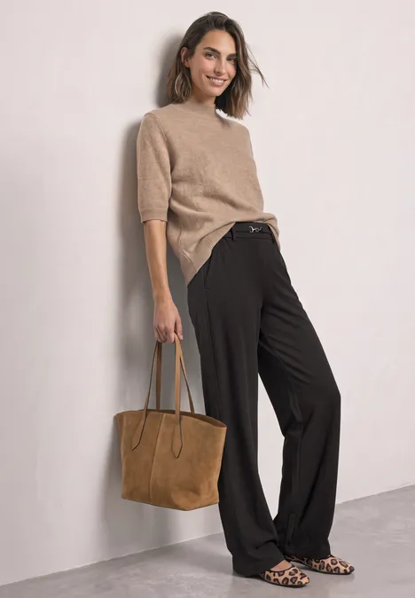 Frau mit beigem Pullover, schwarzen weiten Hosen, Wildleder-Tasche und Leoparden-Print-Ballerinas lehnt an Wand.