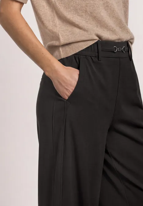 Schwarze, taillierte Hose mit Bügelfalten, beiges Oberteil, Hand steckt in der Seitentasche.