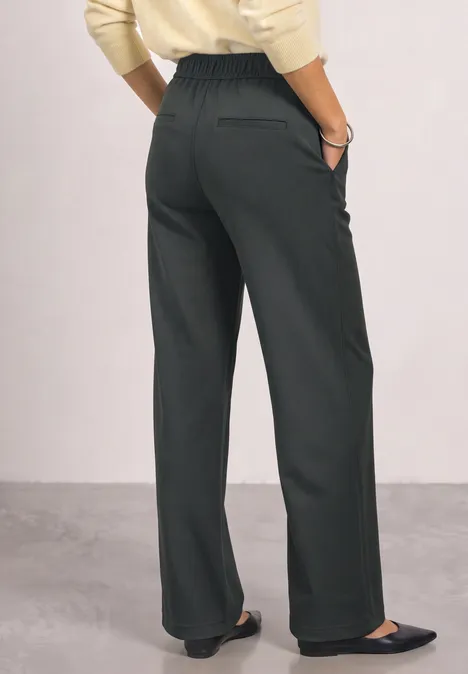 Frau trägt weite, schwarze High-Waist-Hose und schwarze flache Schuhe, Oberteil in Creme, Ansicht von hinten.