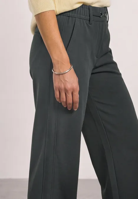 Person trägt dunkelgraue Hose und cremefarbenes Oberteil, Hand mit silbernem Armband sichtbar.