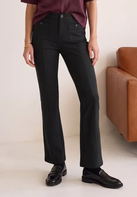 Bootcut Hose mit aufgesetzten Taschen Black Schwarze Hose, schwarze Loafers, violettes Oberteil, stehend vor einem Sofa.