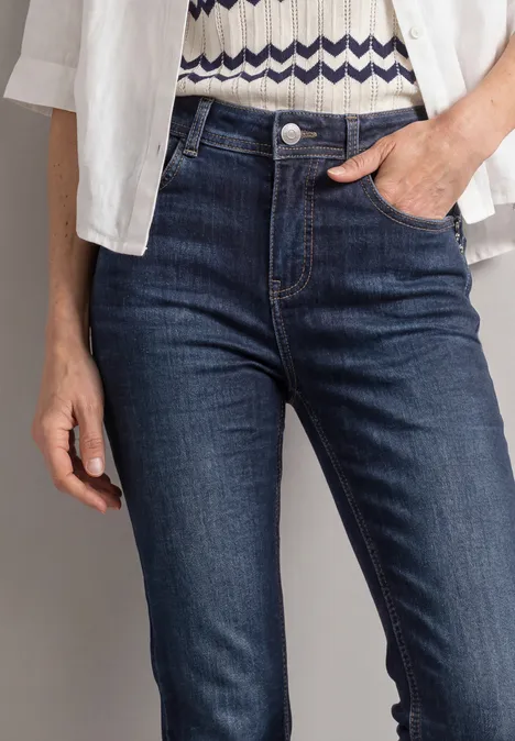 High Waist Bootcut Jeans im Slim Fit Dark Blue Washed Nahaufnahme einer Person in dunkelblauen Jeans mit Hand in der Tasche, weißem Hemd und gemustertem Oberteil.
