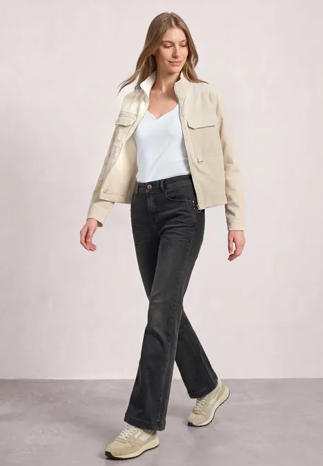 High Waist Bootcut Jeans im Slim Fit Black Washed Frau in cremefarbener Jacke, weißem Top, schwarzen Jeans und Sneakers vor neutralem Hintergrund.