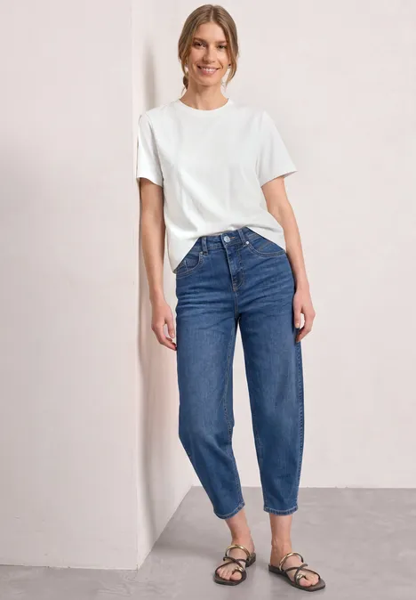 High Waist Barrel Leg Jeans im Loose Fit Medium Blue washed Frau in weißem T-Shirt, locker sitzenden blauen Jeans und schwarzen Sandalen vor schlichtem Hintergrund.