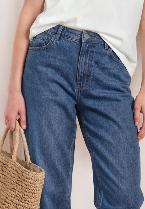 High Waist Barrel Leg Jeans im Loose Fit Medium Blue washed Eine Person trägt blaue Jeans und ein weißes Oberteil, hält eine Strohtasche in der Hand.