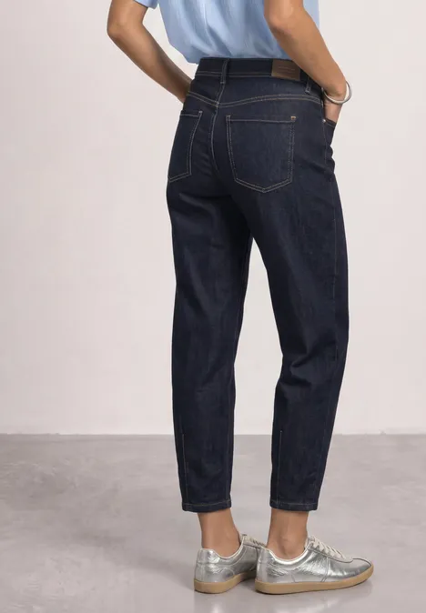 Person trägt hoch geschnittene, dunkelblaue Jeans und silberne Sneakers vor neutralem Hintergrund.