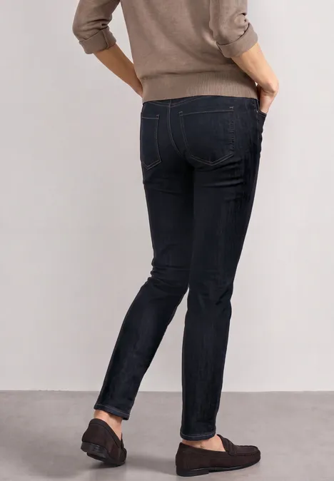 Jean taille moyenne, coupe Slim Leg, style décontracté Deep Rinse Washed Frau in schwarzen Skinny-Jeans und dunklen Slippern seitlich stehend, Hände in den Taschen.