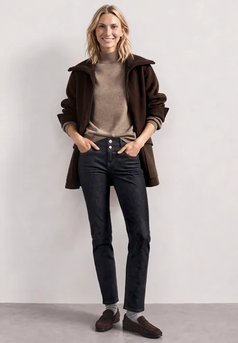 Jean taille moyenne, coupe Slim Leg, style décontracté Deep Rinse Washed Frau in braunem Mantel, Rollkragenpullover, schwarzen Jeans und Loafers vor schlichtem Hintergrund.
