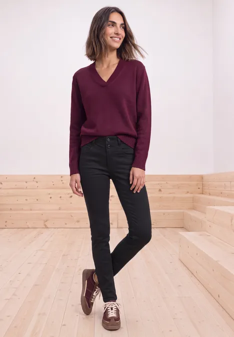 Mid Waist Slim Leg Jeans im Casual Fit Black Clean Washed Frau in weinrotem V-Pullover, schwarzen Jeans und zweifarbigen Schuhen steht lächelnd auf Holzstufen vor weißer Wand.