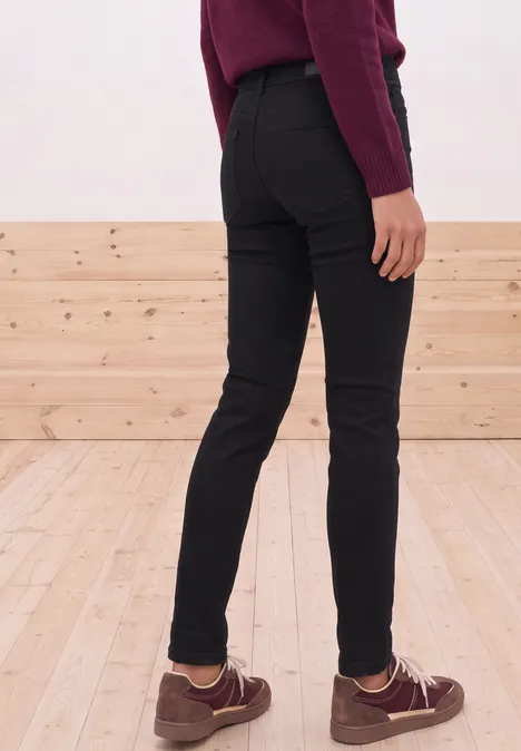 Mid Waist Slim Leg Jeans im Casual Fit Black Clean Washed Eine Person steht in schwarzen, eng anliegenden Hosen und braunen Schuhen vor einer Holzverkleidung.