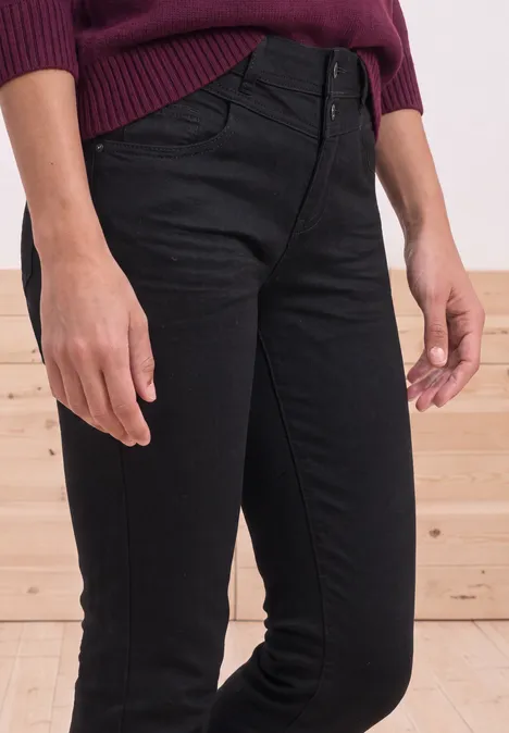 Mid Waist Slim Leg Jeans im Casual Fit Black Clean Washed Schwarze Skinny-Jeans mit hohem Bund und Knopfverschluss, kombiniert mit einem dunkelroten Pullover vor Holzwand.