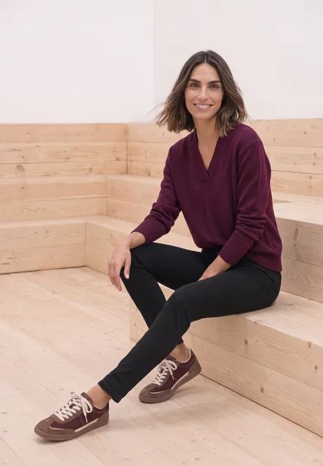 Mid Waist Slim Leg Jeans im Casual Fit Black Clean Washed Eine Frau sitzt auf einer hellen Holzbank, trägt einen burgunderfarbenen Pullover, schwarze Hose und lächelt.