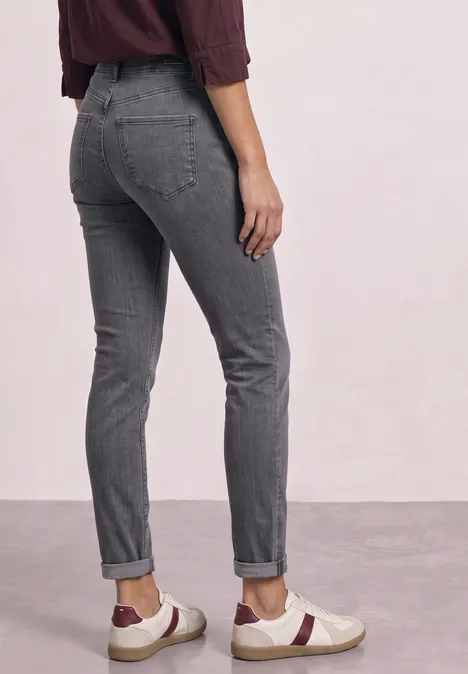 Rugaanzicht van iemand in grijze jeans en witte sneakers met rode streep op een neutrale achtergrond.