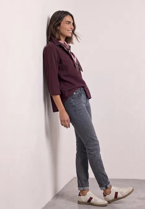 Vrouw lacht, leunt tegen een muur, draagt bordeaux blouse, grijze jeans en comfortabele instappers.