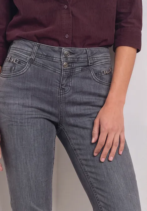 Een vrouw draagt een grijze jeans en een bordeauxrood shirt, alleen het bovenlijf en de hand zijn zichtbaar.