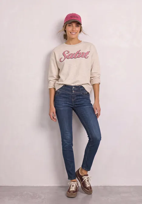 Mid Waist Slim Leg Jeans z ozdobnym detalem Dark Blue Soft Washed Kobieta w dżinsach, beżowej bluzie z napisem 'Salut', różowej czapce i brązowych butach na neutralnym tle.
