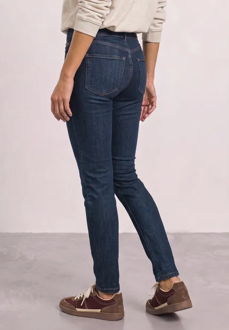 Mid Waist Slim Leg Jeans z ozdobnym detalem Dark Blue Soft Washed Osoba w ciemnych wąskich dżinsach i brązowych butach stoi bokiem na tle jasnej ściany.