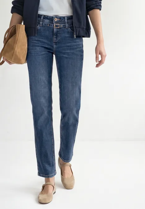 Straight Leg Jeans mit Gürtel Mid Blue Random Washed Frau in blauen Jeans, weißem Shirt, navyblauer Jacke und beigen Schuhen mit Tasche.