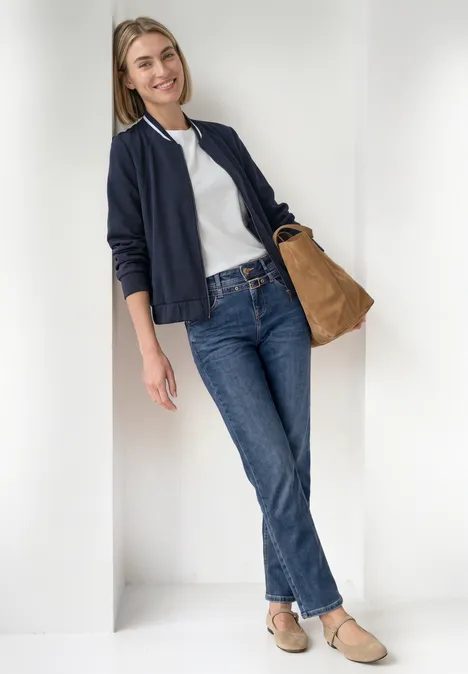 Straight Leg Jeans mit Gürtel Mid Blue Random Washed Eine junge Frau in Jeans, weißem Shirt und Bomberjacke lehnt lächelnd in einem weißen Türrahmen.