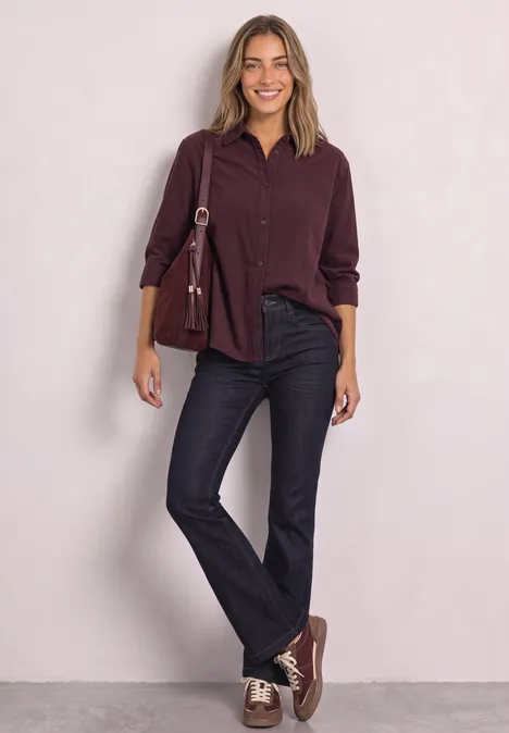 Eine Frau vor hellem Hintergrund trägt weinrotes Hemd, schwarze Jeans, passende Sneaker und eine Umhängetasche.