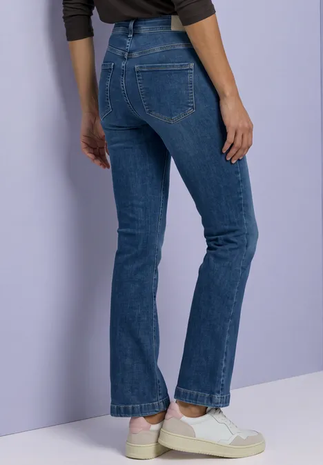 Une personne vue de profil porte un jean bleu et des baskets blanches devant un mur violet clair.