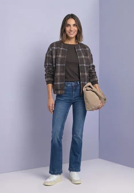 Une femme souriante porte une veste à carreaux, un jean et des baskets blanches, tient un sac beige sur fond violet.