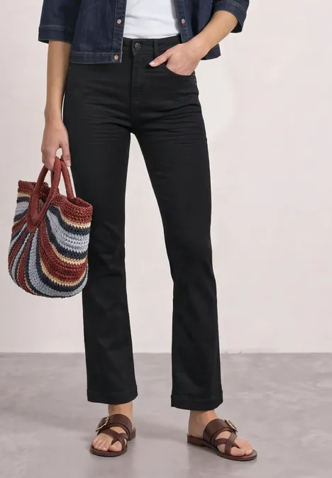 Personne en jean noir, veste en denim, top blanc, sandales marron, tenant un sac rayé multicolore.