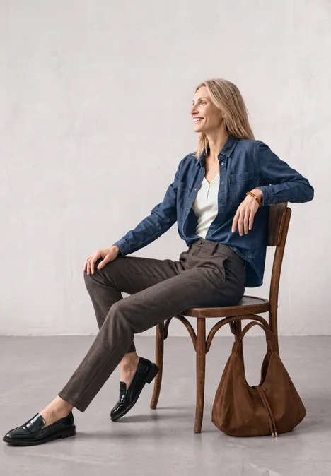 Mid Waist Chino im Casual Fit black coffee Eine Frau sitzt entspannt auf einem Stuhl, trägt elegante Kleidung und blickt lächelnd zur Seite.