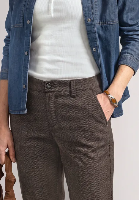 Mid Waist Chino im Casual Fit black coffee Frau trägt weiße Bluse, offene Jeansjacke und braune Hose, Hand in Tasche mit Armbanduhr.