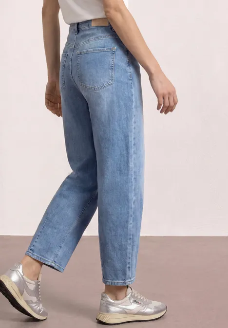 Seitenansicht einer Person in hellblauen, locker sitzenden Jeans und silbernen Sneakern vor neutralem Hintergrund.