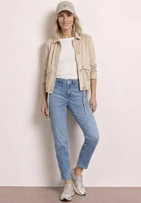 Frau trägt beige Jacke, weißes Shirt, hellblaue Jeans, silberne Sneaker und Basecap.