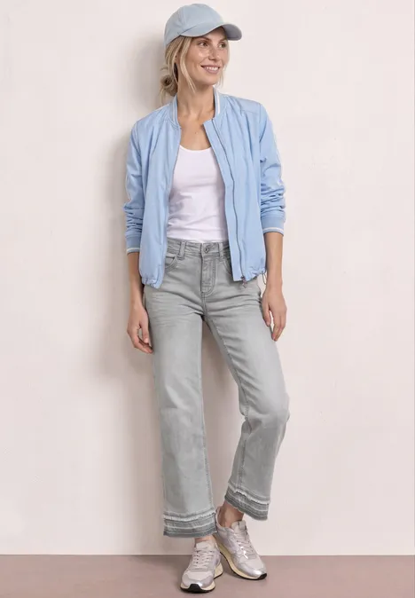 Person trägt hellblaue Bomberjacke, weißes Shirt, graue Cropped-Jeans, Sneakers und Cap vor neutralem Hintergrund.