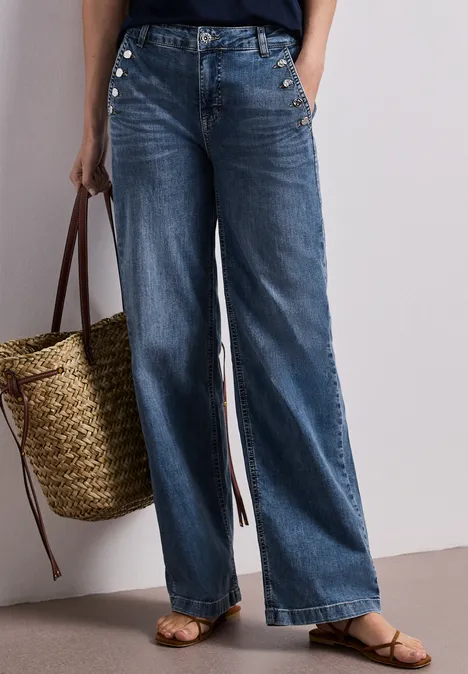 Een vrouw draagt wijdvallende jeans, sandalen en houdt een gevlochten strotas vast.