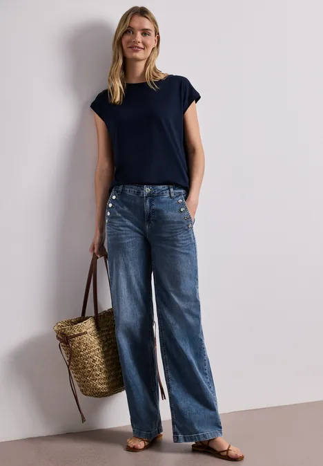 Vrouw in donkerblauw T-shirt, wijde jeans, platte sandalen, met een gevlochten rieten tas.
