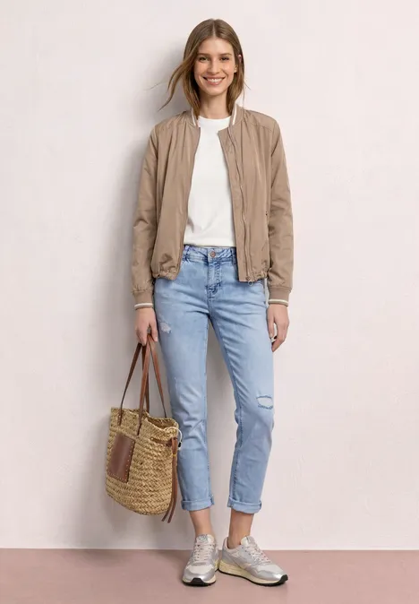 Dame in beiger Jacke, weißem Shirt, blauen Jeans und Sneakers mit geflochtener Tasche vor hellem Hintergrund.