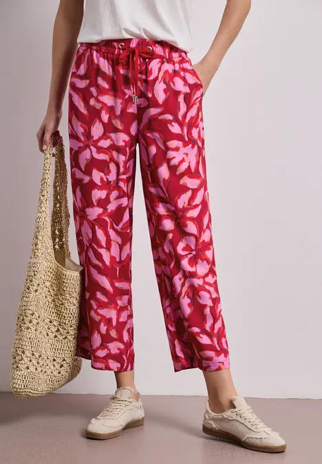 Frau trägt rote, locker sitzende Blumenhose, weiße Sneaker und eine Strohtasche.