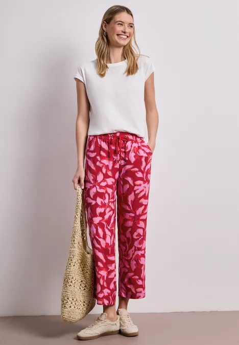 Frau in weißem Oberteil und roten, floralen Culotte-Hosen, trägt Basttasche und Sandalen vor neutralem Hintergrund.