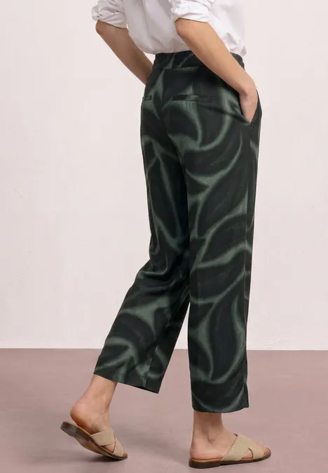 Person trägt weite, dunkelgrün gemusterte Hose mit abstrakten Wirbeln und flache Sandalen.
