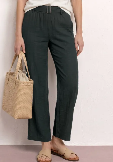 Frau trägt hohe, weite Leinenhose mit Gürtel, hält Korbtasche und trägt flache Sandalen.