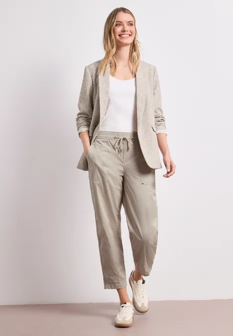Eine Frau trägt lässige, beige Leinenhose, weiße Bluse und Blazer, dazu weiße Sneaker.