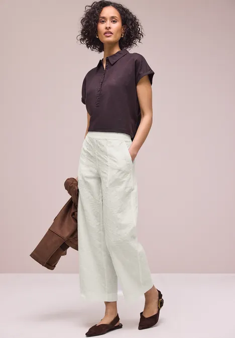 Vrouw in witte, wijdvallende broek en donker poloshirt, met een bruine jas en bruine loafers.