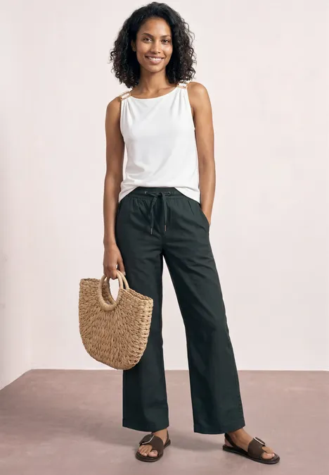 Frau in weißem Tanktop und schwarzen, weiten Hosen trägt Strohtasche und flache Sandalen.