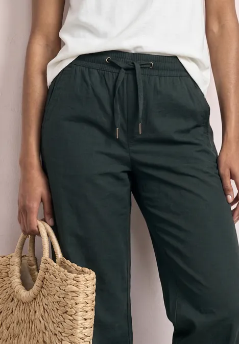 Dunkelgrüne Leinenhose mit Tunnelzug, seitlichen Taschen und Strohtasche in der Hand.