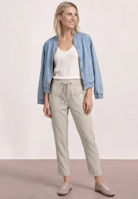 Femme en top blanc, pantalon beige et veste en jean bleu clair, posant devant un fond neutre