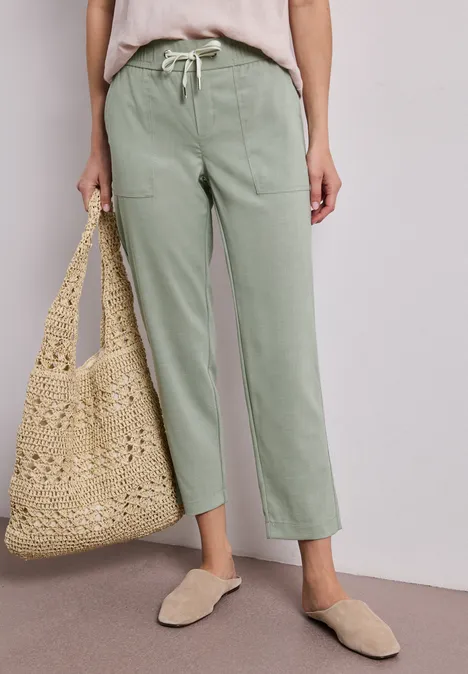 Person trägt hellgrüne, gerade geschnittene Hose, beiges Top, Basttasche und bequeme Slipper.