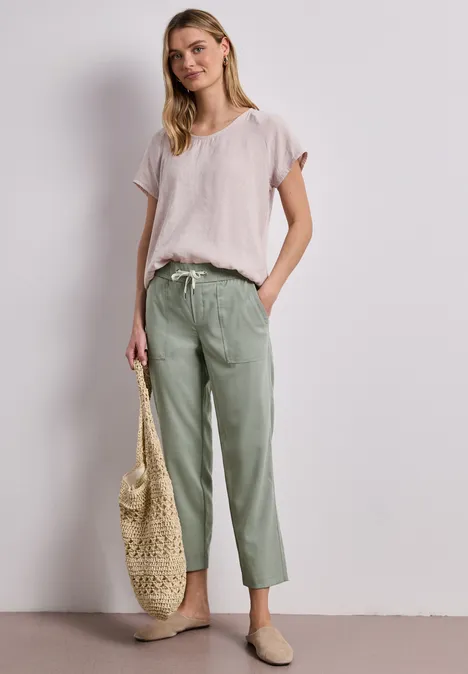 Frau in lockerem hellrosa T-Shirt und grüner Stoffhose, trägt Basttasche und Slipper vor neutralem Hintergrund