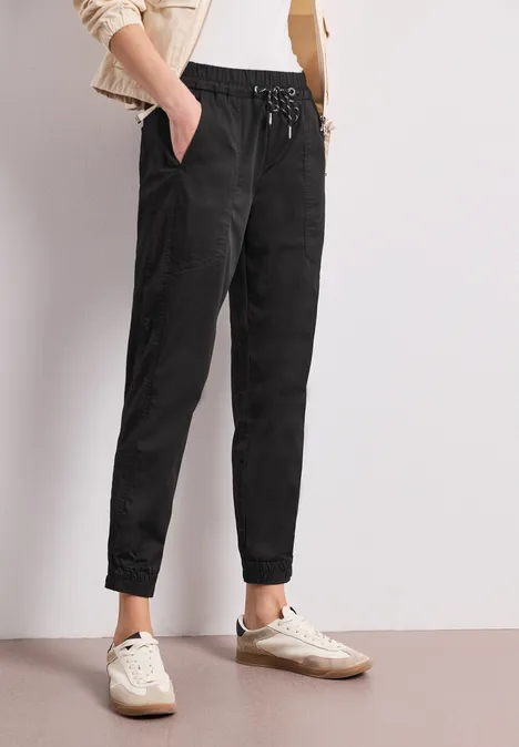 Person in schwarzer, lässiger Jogginghose mit Gummizug, weißen Sneakern vor schlichtem hellem Hintergrund.