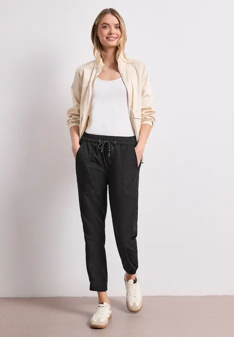 Person mit beigem Hoodie, weißem Top, schwarzen Jogginghosen und weißen Sneakern vor hellem Hintergrund.